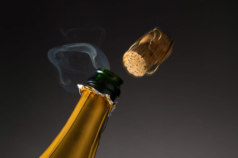 The neck of a. bottle of champagne, cork popping but gently with a plume of smoke.